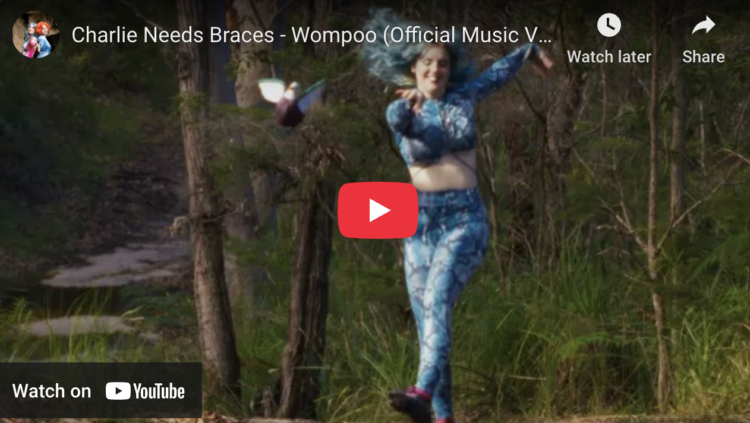 Screenshot of Charlie Needs Braces - Wompoo (Official Music Video) on YouTube features a woman with blue hair and in a blue lycra top and matching leggings skip through the woods
